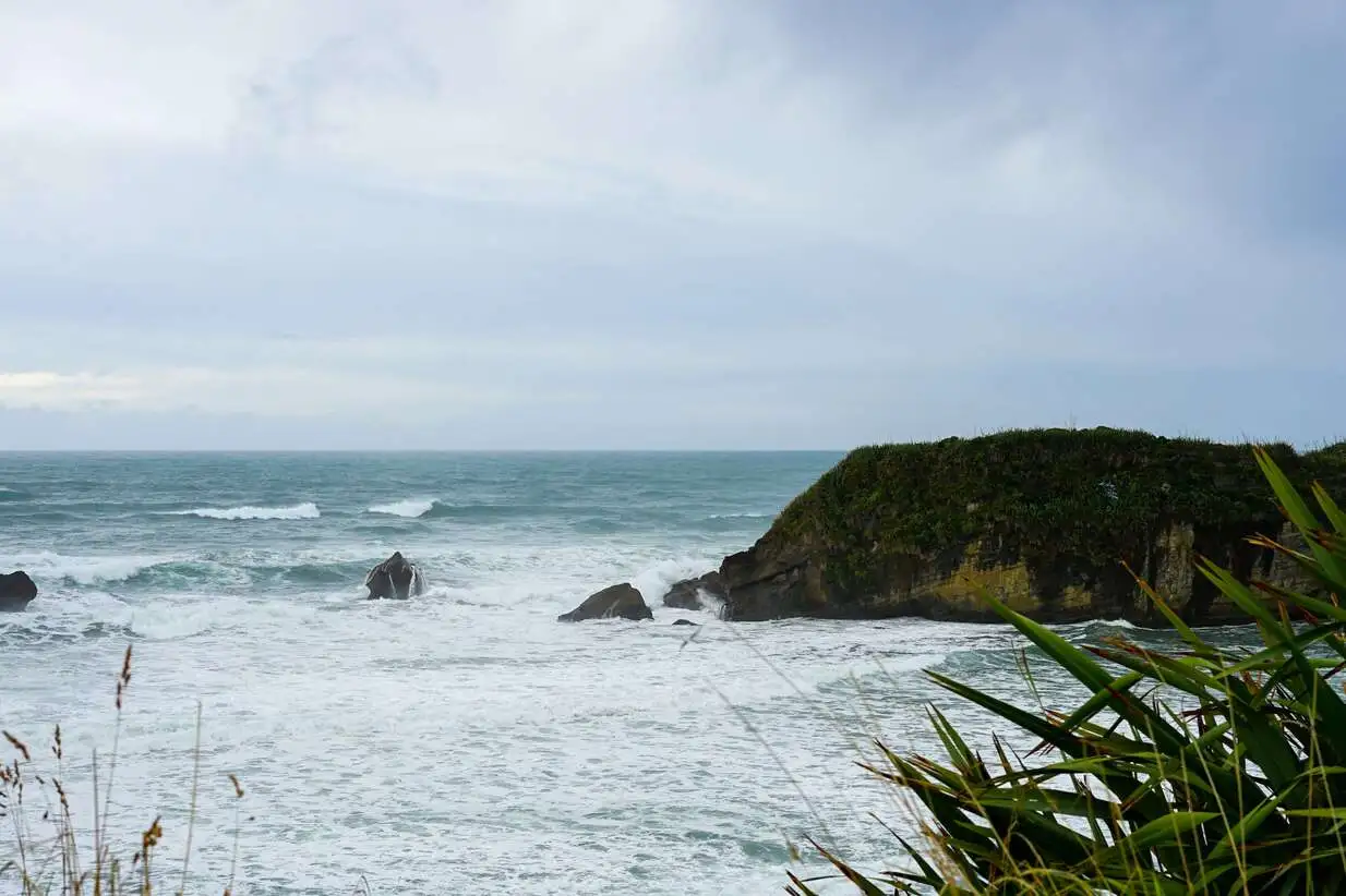 A scenic view of the ocean with waves crashing against rocky outcrops and a small, green-covered island under a cloudy sky.